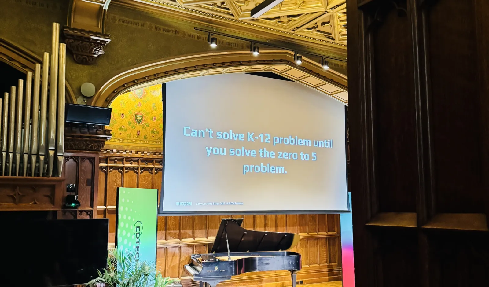A slide on a large screen in a wood-paneled hall with a pipe organ and a grand piano reads: Can’t solve K-12 problem until you solve the zero to 5 problem.
